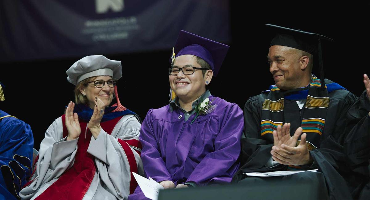 A student sitting between two administrators at commencement A student sitting between two administrators at commencement
